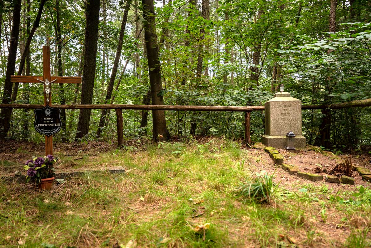 Fotografia współczesna cmentarza rodzinnego rodziny von Finkenstein, dawnych właścicieli zamku. Fotografia przedstawia ogrodzony cmentarz rodzinny. Po prawej stronie znajduje się odnowiony piaskowy sześcian na nieco szerszym postumencie. To bryła nagrobka. Przed nim z kamieni ułożono kształt symbolizujący powierzchnię grobu przed jego dewastacją. Po lewej stronie fotografii znajduje się prosty, drewniany krzyż z figurą Jezusa i tablicą informującą o poświęceniu tego miejsca pamięci rodziny von Finkenstein. Cmentarz ulokowany jest w lesie, porasta go trawa.